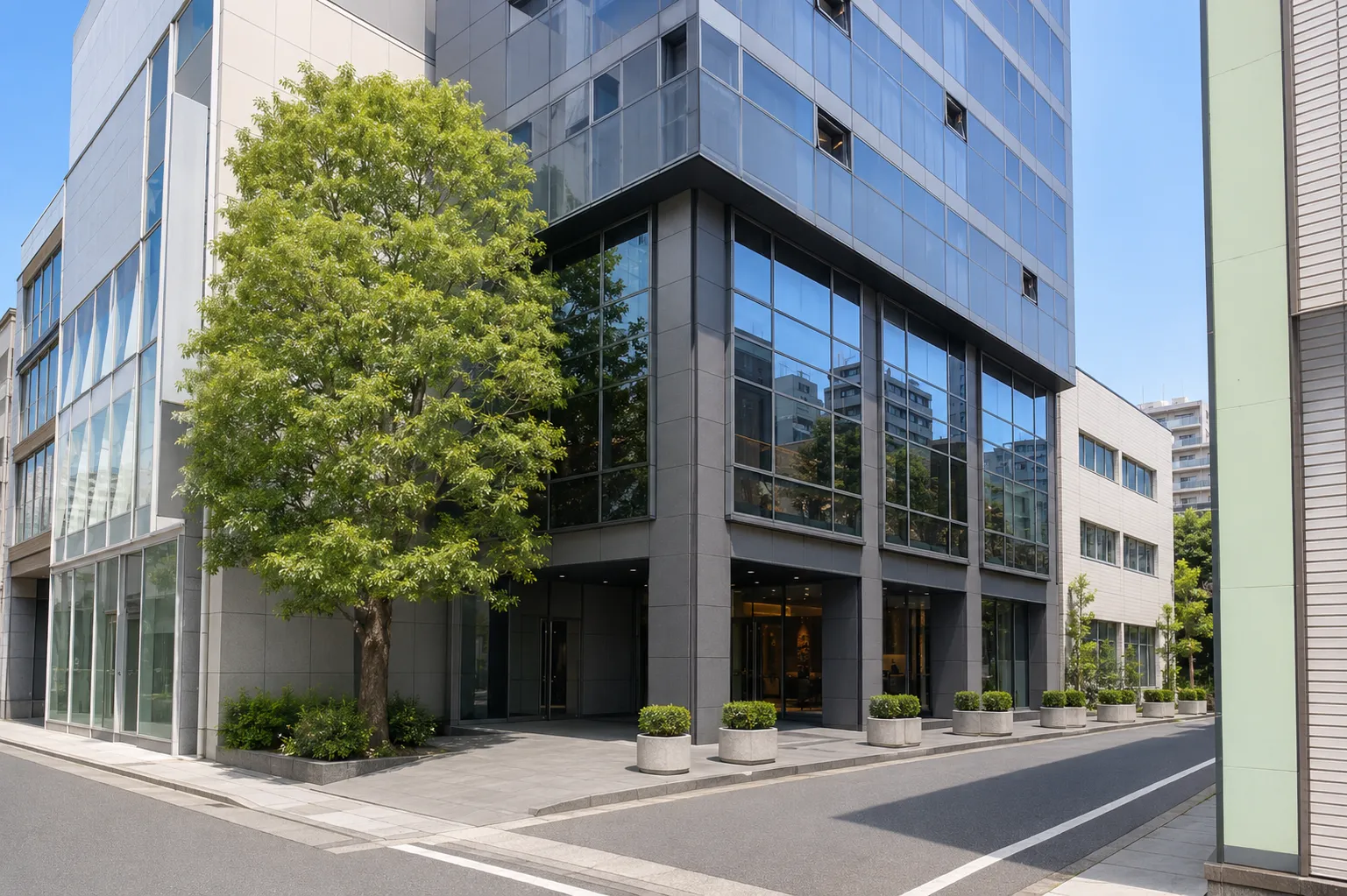 東京オフィスが入る建物の外観。角地に建ち、1階から2階までが濃いグレーの石張り、その上がガラス張りのオフィスビル。歩道に面した1階の中央に奥まったガラスのエントランスがあり、その前に円柱形の植栽プランターが等間隔に並ぶ。左手前に大きな街路樹が立つ。